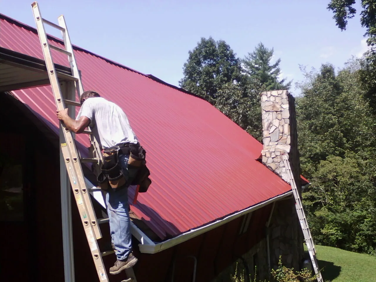Roofer on a ladder beside a red metal roof and stone chimney — Soffit & Fascia Repair by Roofing Brothers in Hyndman